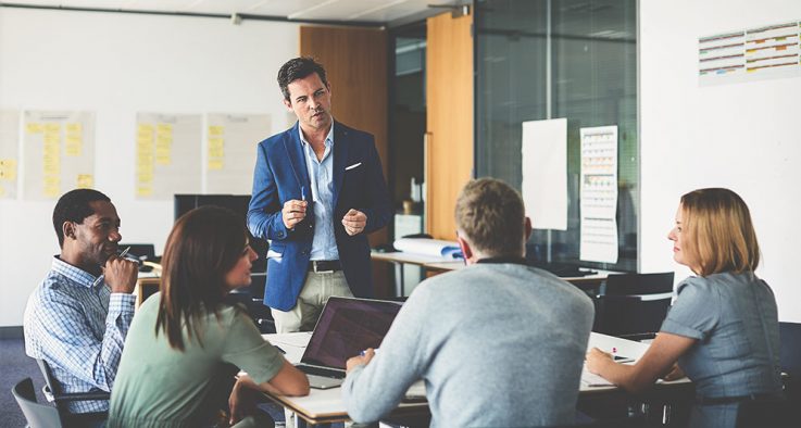 Image of colleagues in a meeting with one person standing up front making a point while holding a pen.